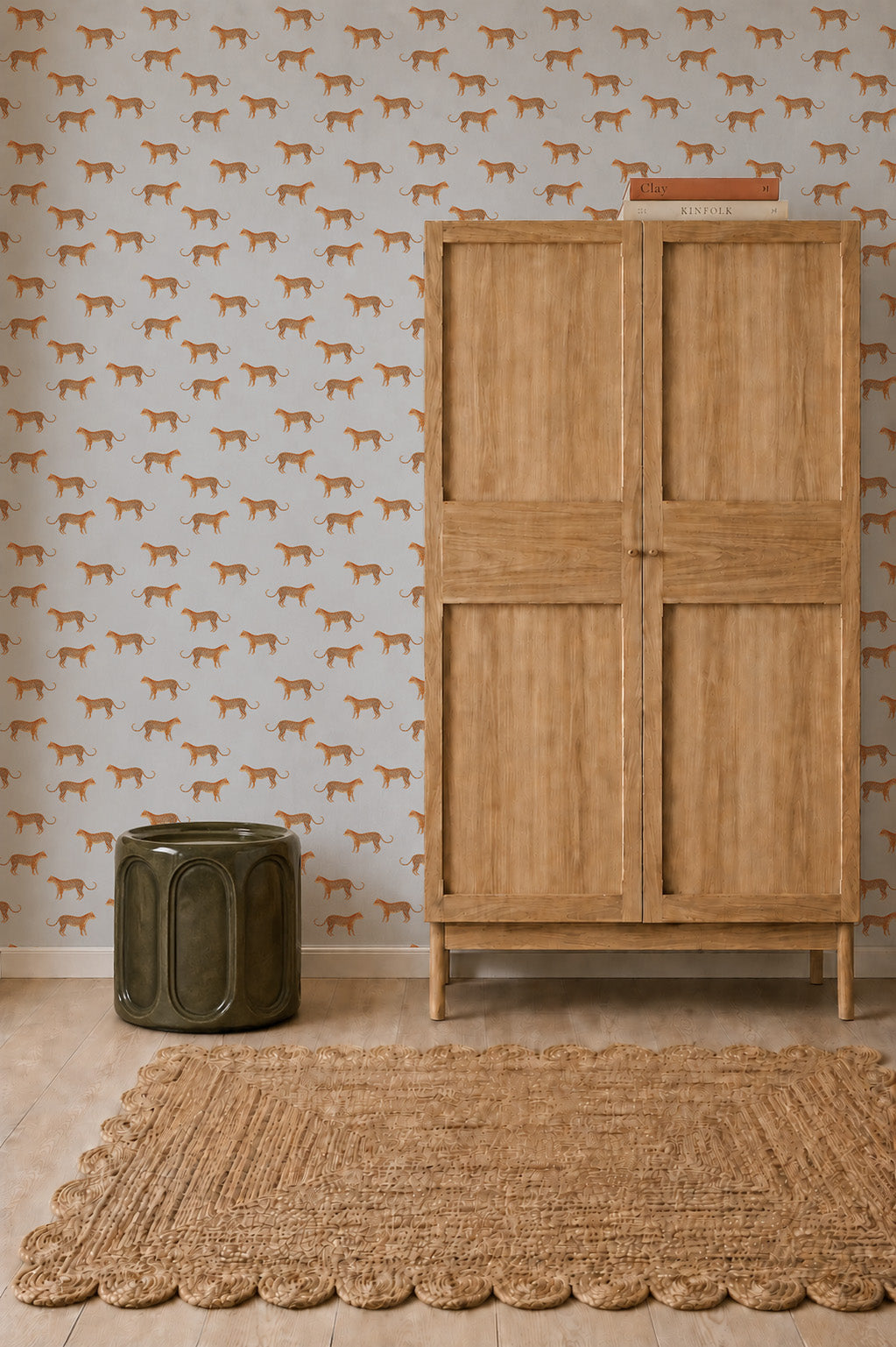 A wooden cabinet stands next to a green metal stool on a woven rug. The wall behind features Goldies Cheetah Wallpaper in blue, bringing a lively touch to the scene with its playful pattern. Two books rest on top of the cabinet.