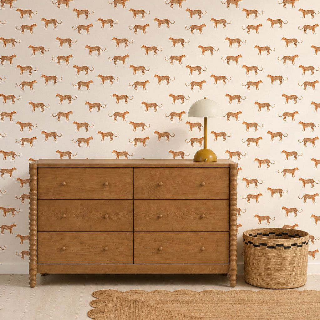 A wooden dresser sits against a wall adorned with Goldies Cheetah Wallpaper in cream, perfect for a kids room. Nearby, a small lamp and woven basket add charm, while a scalloped rug completes the cozy space.