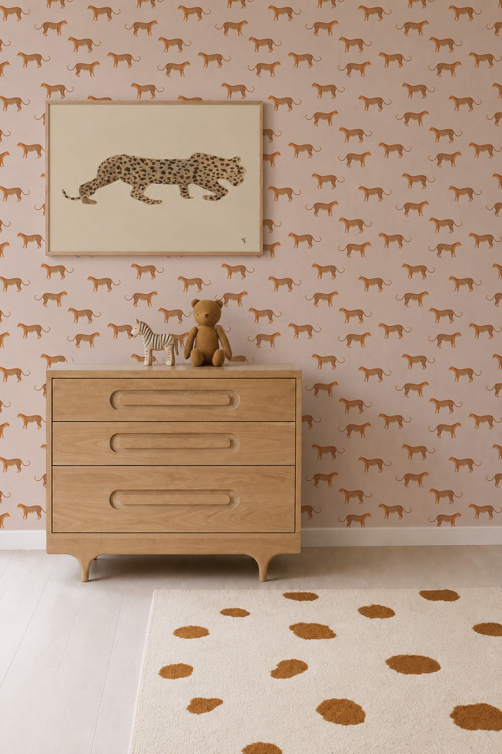 A dresser with a zebra and teddy bear sits below a framed cheetah print on a wall covered in Goldies Cheetah Wallpaper, Pink. A beige rug with brown spots is on the light wood floor.