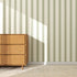 A wooden dresser with three drawers stands on a woven rug against a wall covered in Scallop Stripe Wallpaper, Green. Sunlight casts a rectangular shadow over the wallpaper’s vertical, scalloped green stripes.