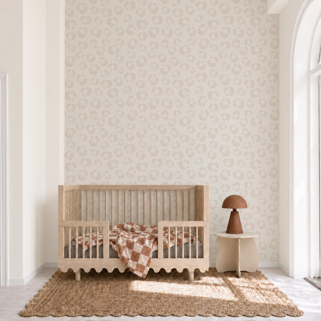 A minimalist nursery featuring a wooden crib, brown and white checkered blanket, small table with a brown lamp, jute rug, and the Hallie Leopard Print Wallpaper Sample in Light Pink showcases exceptional wallpaper quality. The room sits next to a tall arched window letting in natural light.