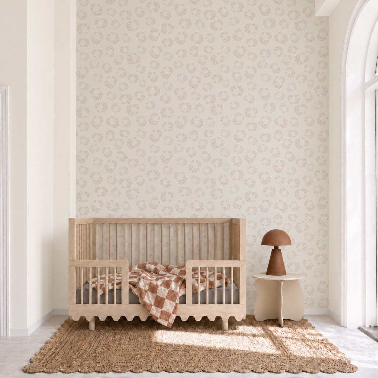 A minimalist nursery featuring a wooden crib, brown and white checkered blanket, small table with a brown lamp, jute rug, and the Hallie Leopard Print Wallpaper Sample in Light Pink showcases exceptional wallpaper quality. The room sits next to a tall arched window letting in natural light.