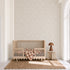 A minimalist nursery featuring a wooden crib, brown and white checkered blanket, small table with a brown lamp, jute rug, and the Hallie Leopard Print Wallpaper Sample in Light Pink showcases exceptional wallpaper quality. The room sits next to a tall arched window letting in natural light.