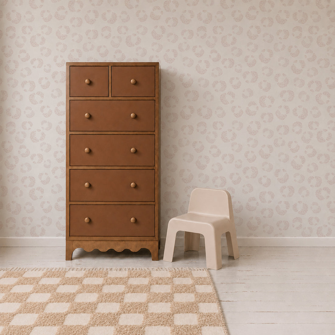 A brown wooden dresser with six drawers stands beside a small beige plastic chair. The room features Hallie Leopard Print Wallpaper Sample in light pink, showcasing excellent wallpaper quality, along with a white floor and a beige checkered rug.