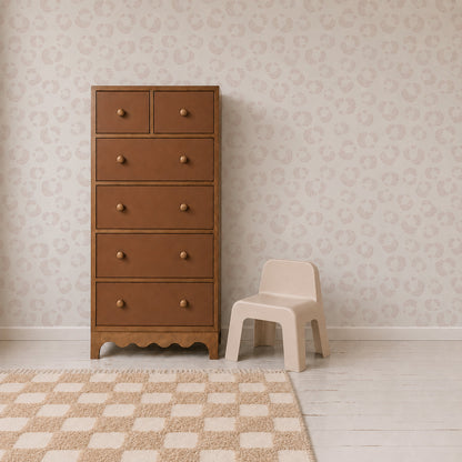 A brown wooden dresser with six drawers stands beside a small beige plastic chair. The room features Hallie Leopard Print Wallpaper Sample in light pink, showcasing excellent wallpaper quality, along with a white floor and a beige checkered rug.