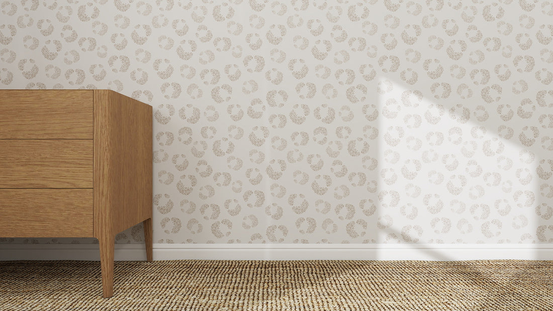 A wooden dresser with three drawers stands on a textured carpet beside a wall decorated with Hallie Leopard Print Wallpaper Repeat Pattern in beige, as sunlight casts a triangular shadow on the wall.