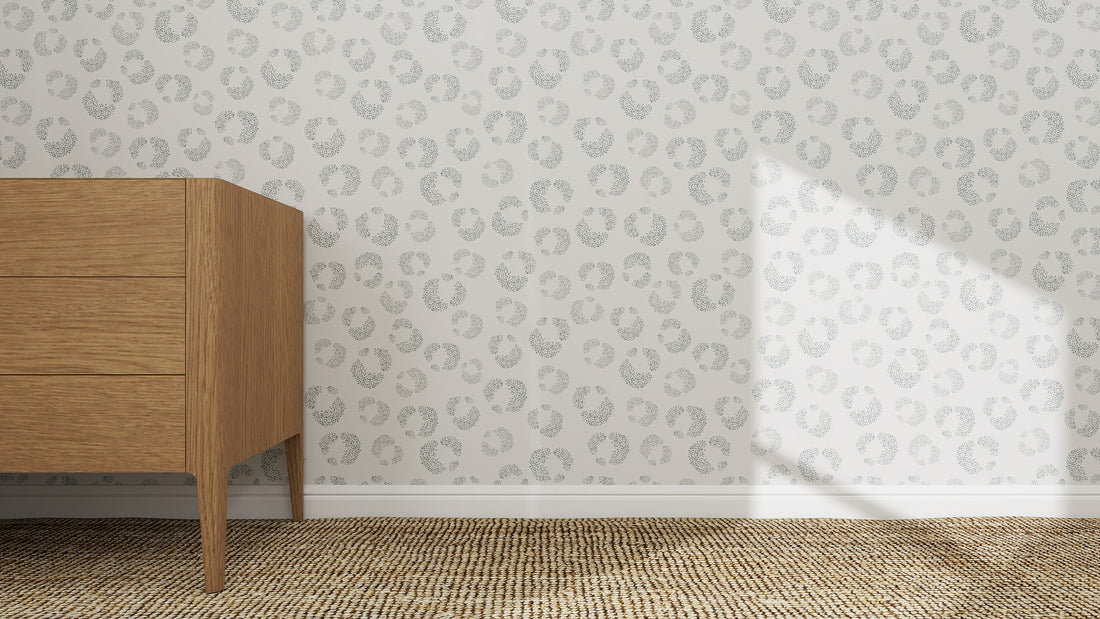A wooden dresser stands on a woven carpet against a white wall decorated with Hallie Leopard Print Wallpaper Repeat Pattern in light blue. Sunlight casts a triangle-shaped shadow across the playful wallpaper.