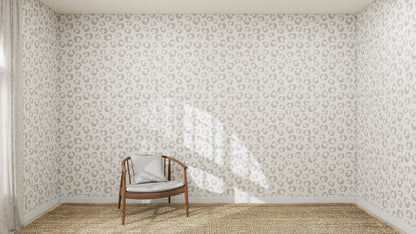 A wooden chair with a gray cushion sits in the corner of a room, accented by Hallie Leopard Print Wallpaper in charcoal, a woven rug floor, and sunlight streaming through the window.