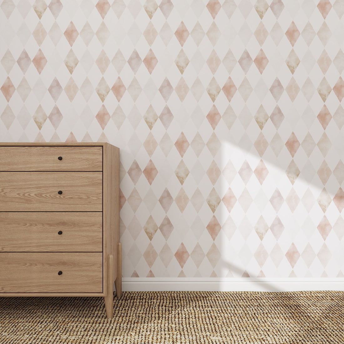 A light wooden dresser with four drawers stands on a woven rug against a wall covered in Harlequin Watercolour Wallpaper Repeat Pattern | Pink, featuring pastel diamonds. Sunlight casts playful shadows on the wallpaper from the right side.