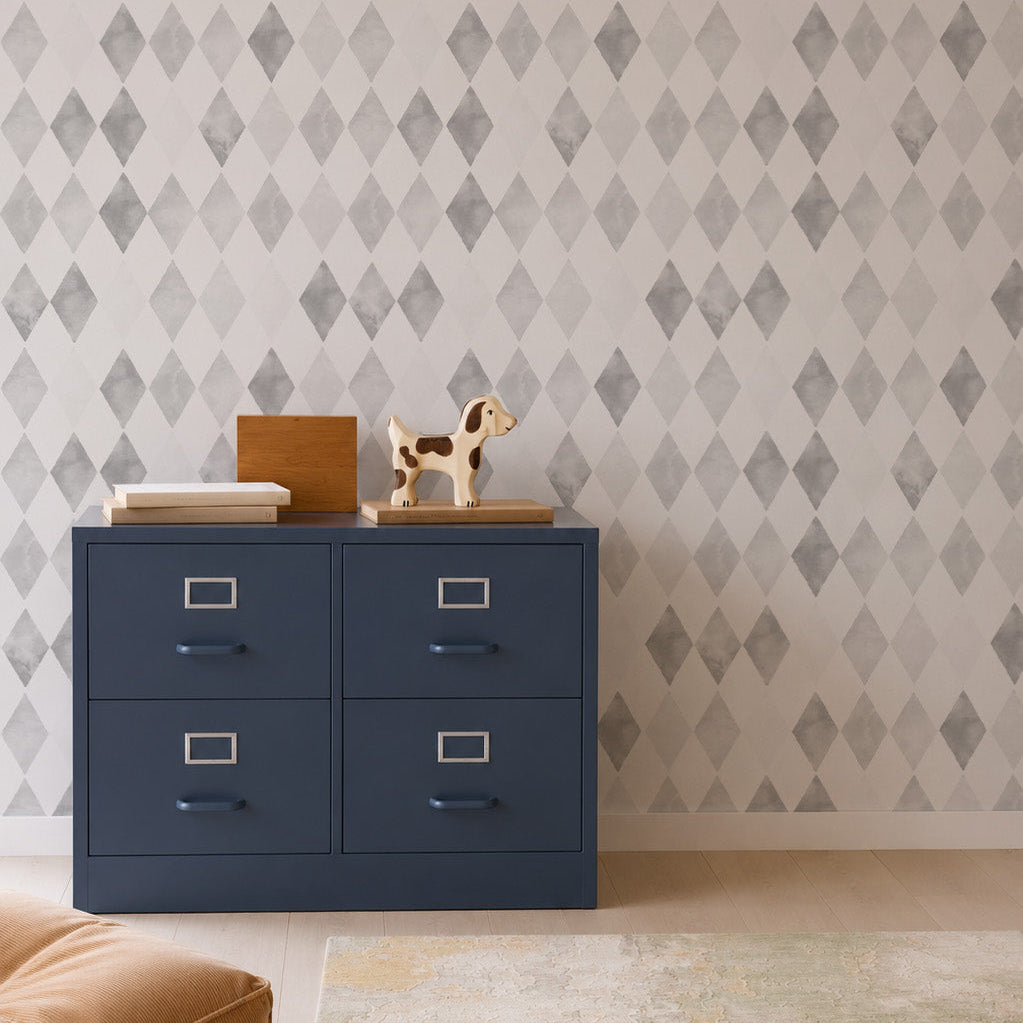 A blue four-drawer cabinet with books and a toy dog on top sits against a playful pattern of Harlequin Watercolour Wallpaper, Grey. A tan floor cushion and a light-colored rug are in the foreground.