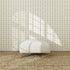 A white ottoman with wooden legs rests on a woven rug before the Jolly Jester Wallpaper Sample in sage green, as sunlight pours in and window-shaped shadows highlight the wallpaper’s color and pattern.