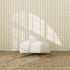 A white, round ottoman sits on a textured woven rug in front of the Jolly Jester Wallpaper Repeat Yellow sample. Sunlight streams through a window, casting geometric shadows that highlight the wallpaper’s quality.