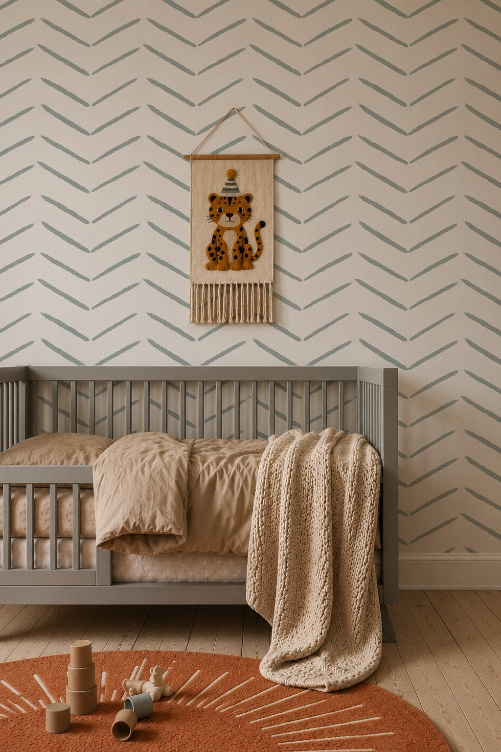 A cozy nursery with a gray crib, beige bedding, a knitted blanket, wooden toys on an orange rug, and the wall adorned with Herringbone Wallpaper in blue plus a hanging tapestry featuring a leopard illustration.