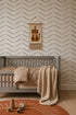 A cozy nursery with a gray crib, beige bedding, a knitted blanket, wooden toys on an orange rug, and the wall adorned with Herringbone Wallpaper in blue plus a hanging tapestry featuring a leopard illustration.
