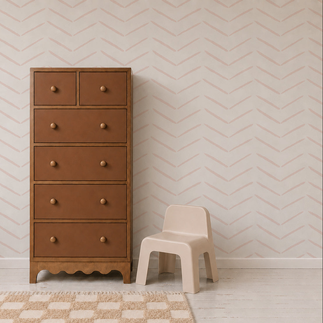 A tall brown dresser with round knobs stands beside a small beige children’s chair on a light wood floor, set against a white wall decorated with Herringbone Wallpaper in Rose, with a beige patterned rug in front.