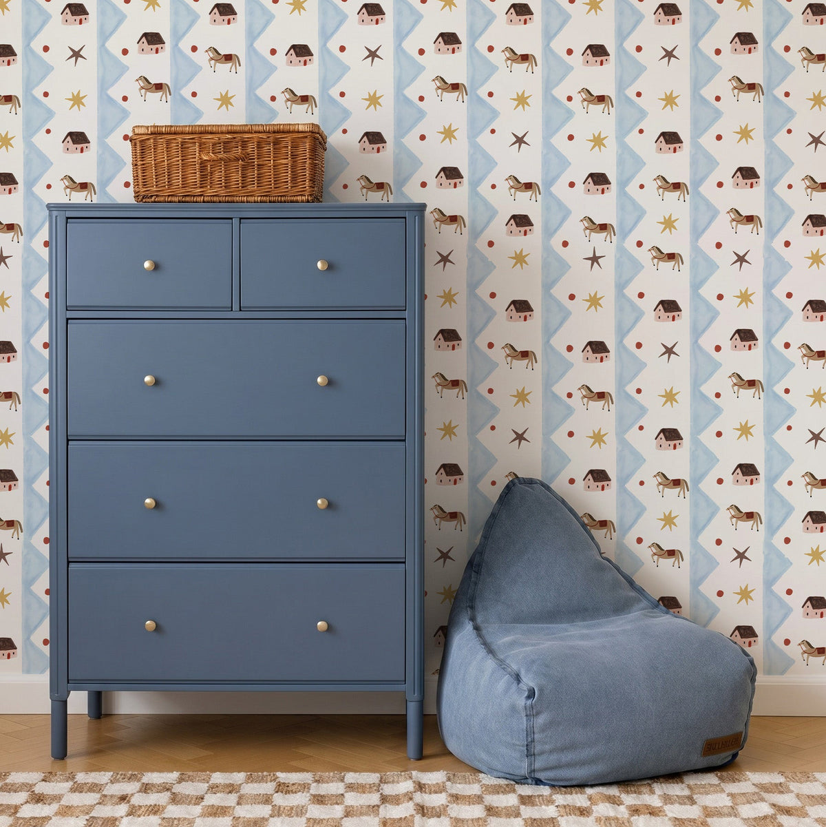A blue dresser with five drawers stands beside a blue bean bag chair. The wall is decorated with Paddock Parade Wallpaper Sample, featuring playful houses, animals, and stars that add vibrant color to the room. A wicker basket sits on top of the dresser, and a checkered rug is on the floor.