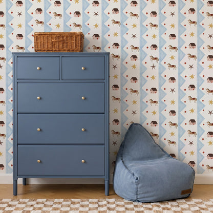A blue dresser with five drawers stands beside a blue bean bag chair. The wall is decorated with Paddock Parade Wallpaper Sample, featuring playful houses, animals, and stars that add vibrant color to the room. A wicker basket sits on top of the dresser, and a checkered rug is on the floor.