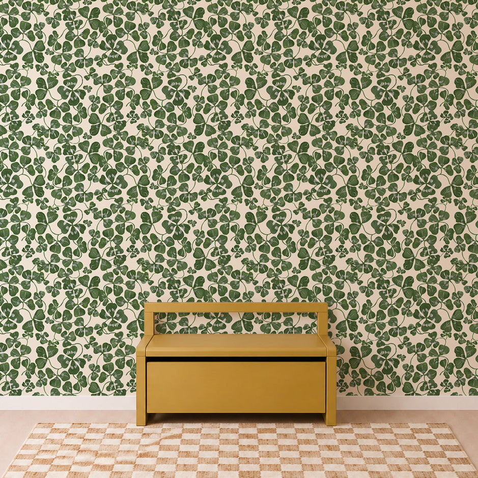 A yellow bench sits against a wall adorned with the “How Lucky We Are Clovers Wallpaper,” featuring lush green leaves. The floor showcases a beige and white checkerboard rug, enhancing the simple and well-lit setting. With its minimalist, modern style, this space could easily serve as inviting nursery wallpaper inspiration.