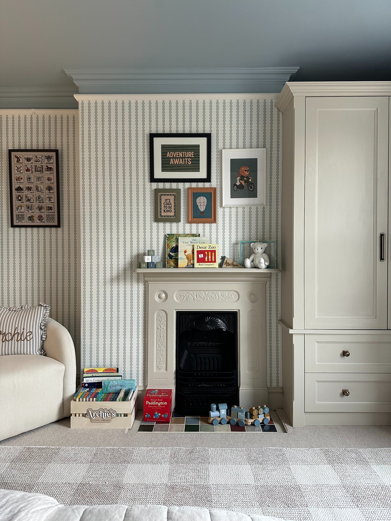 A cozy children’s room features Little Abacus Stripe Blue and Green nursery wallpaper, a white fireplace with framed art, books, toys, a teddy bear on the mantel, plus a cream wardrobe and cushioned sofa.