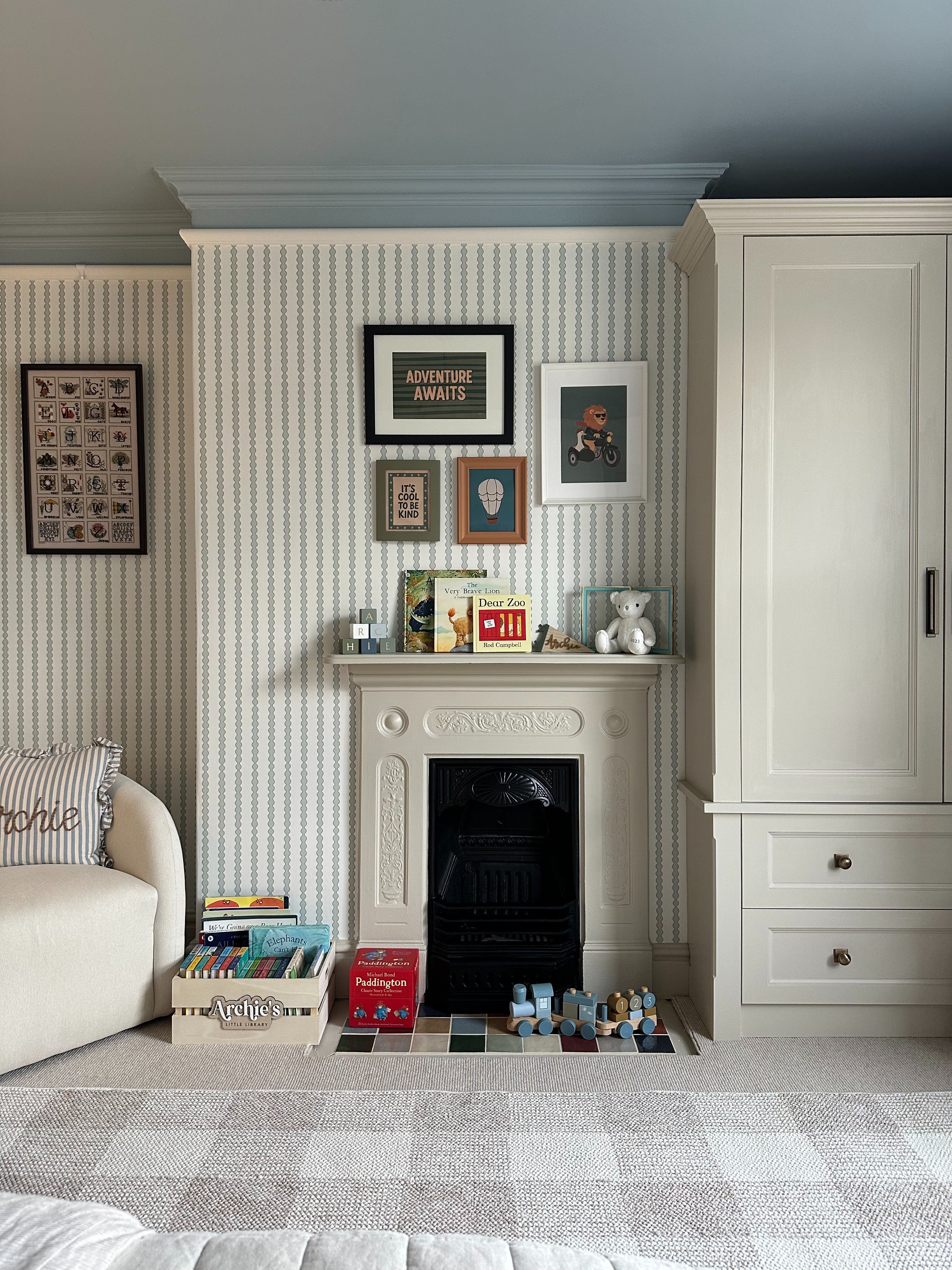 A cozy children’s room features Little Abacus Stripe Blue and Green nursery wallpaper, a white fireplace with framed art, books, toys, a teddy bear on the mantel, plus a cream wardrobe and cushioned sofa.