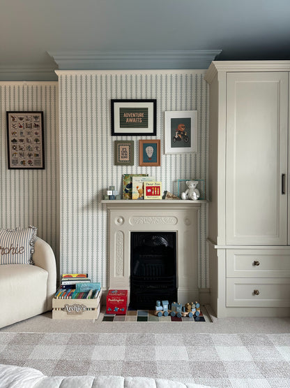 A cozy children’s room features Little Abacus Stripe Blue and Green nursery wallpaper, a white fireplace with framed art, books, toys, a teddy bear on the mantel, plus a cream wardrobe and cushioned sofa.