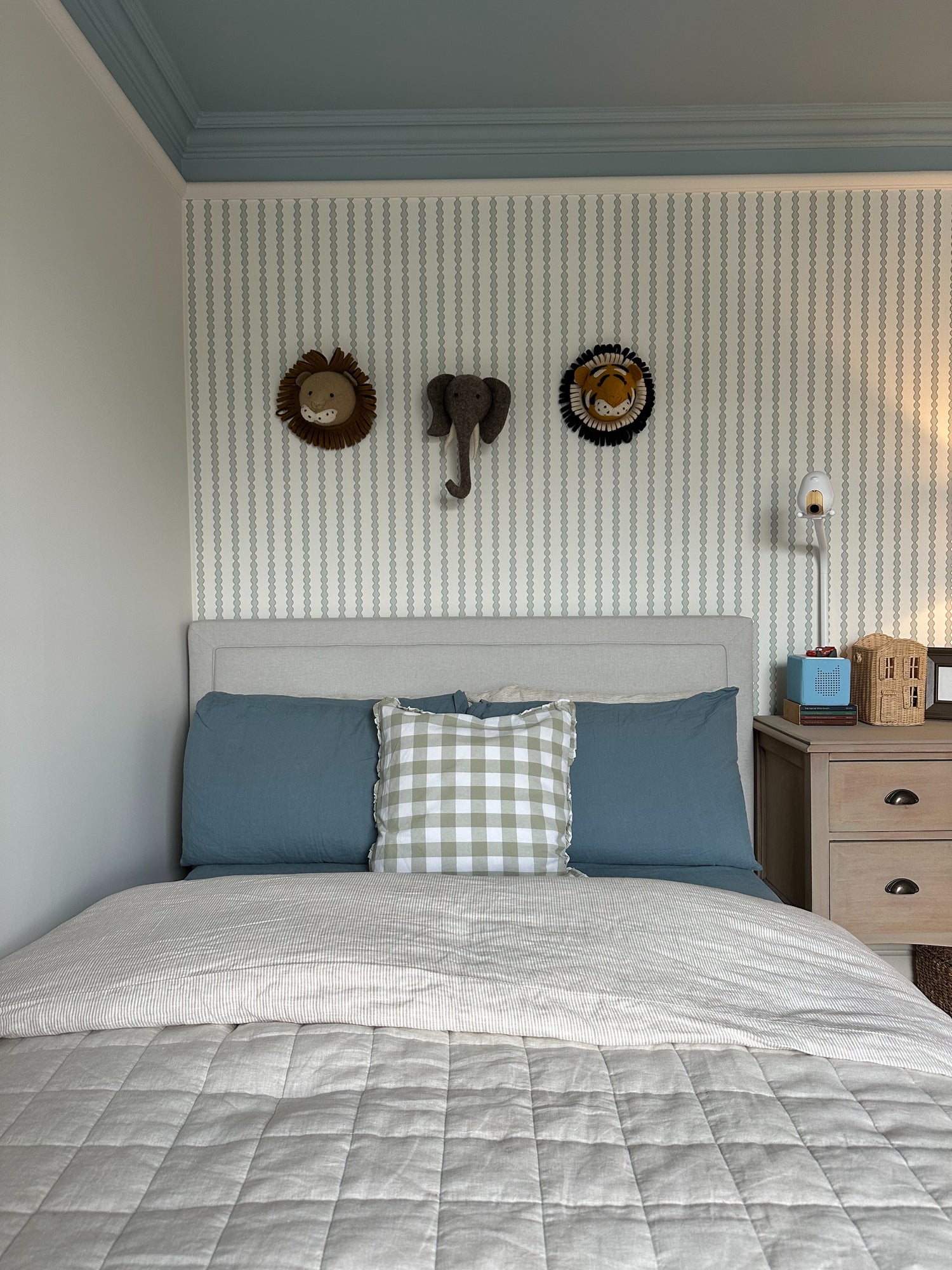 A neatly made bed with blue and gray bedding, a plaid pillow, and a beige headboard sits against a wall decorated with Little Abacus Stripe Blue and Green wallpaper. Above the bed are three animal decorations: two lions and an elephant.