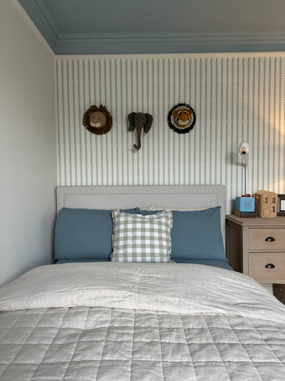 A neatly made bed with blue and gray bedding, a plaid pillow, and a beige headboard sits against a wall decorated with Little Abacus Stripe Blue and Green wallpaper. Above the bed are three animal decorations: two lions and an elephant.