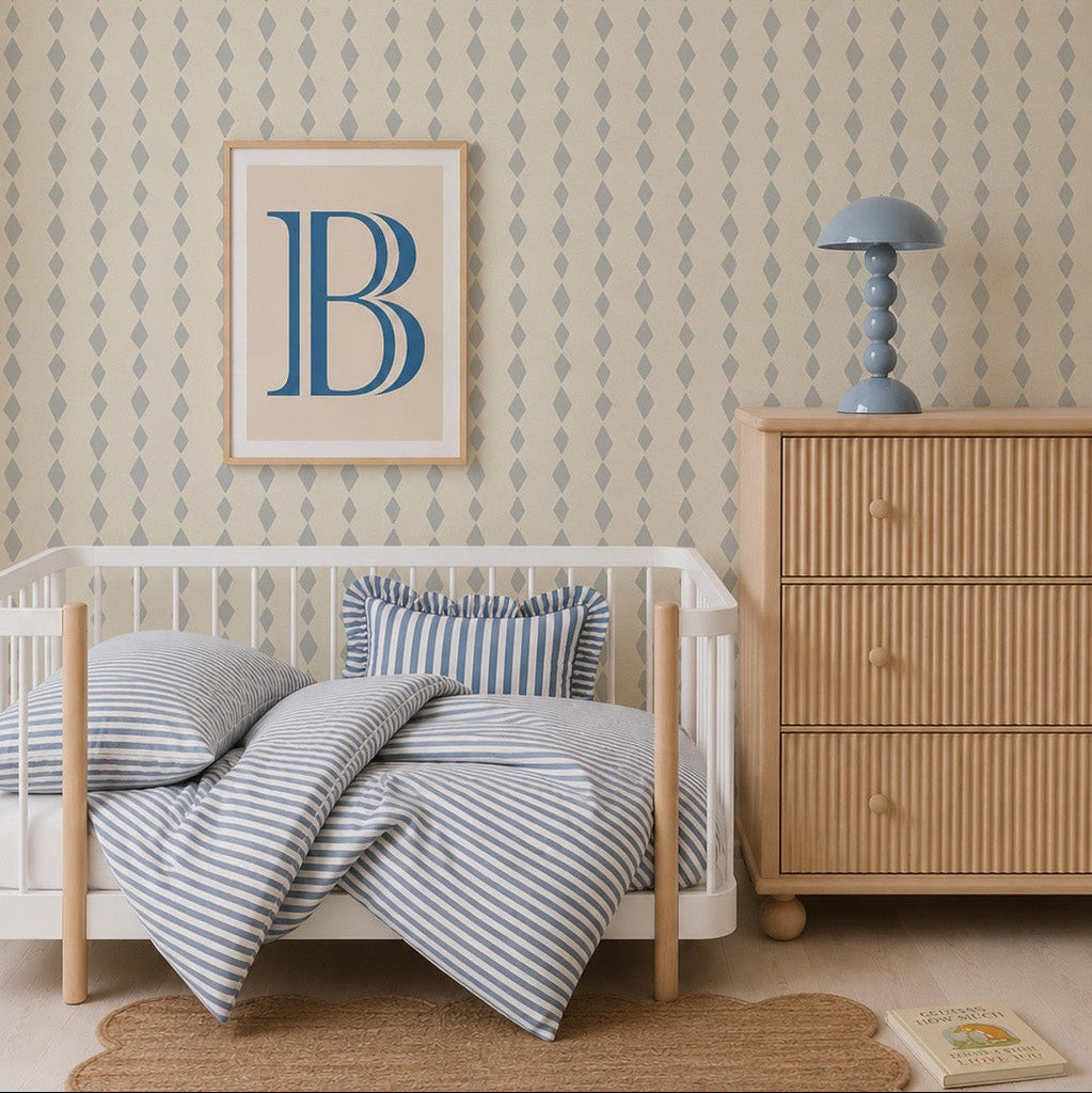 A cozy nursery with a wooden crib, striped blue and white bedding, a light wood dresser, a blue lamp, a framed letter B on the wall, and Jolly Jester Wallpaper, Blue featuring a playful harlequin pattern.