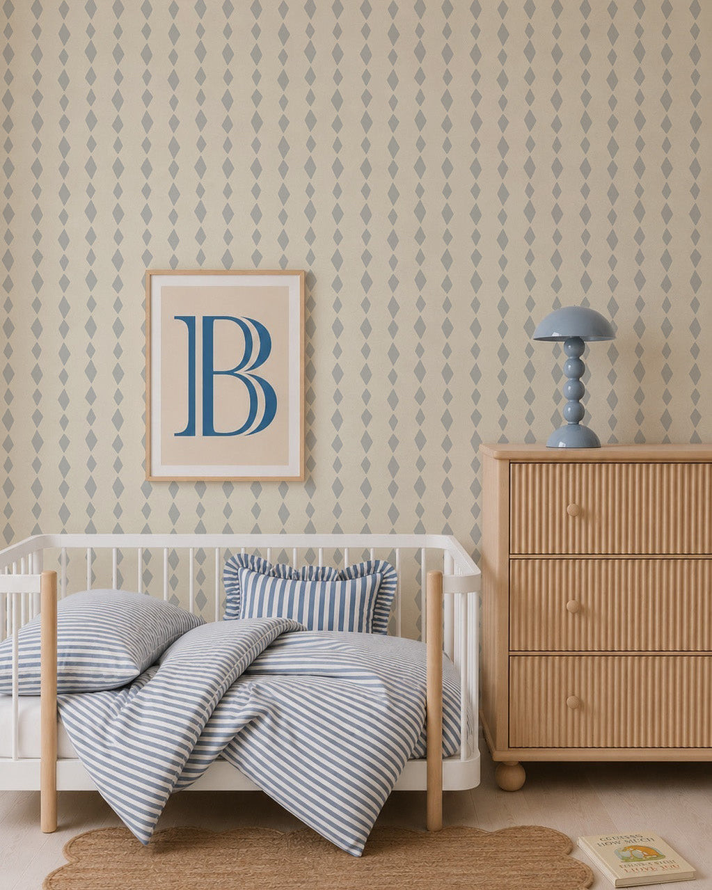 A cozy nursery with a wooden crib, striped blue and white bedding, a light wood dresser, a blue lamp, a framed letter B on the wall, and Jolly Jester Wallpaper, Blue featuring a playful harlequin pattern.