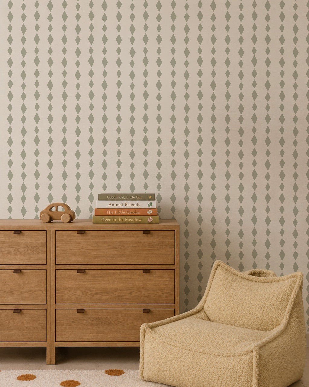 A neutral-toned room with a wooden dresser, beige textured armchair, and stacked books. The wall features Jolly Jester Wallpaper in sage green, adding playful elegance perfect for kids&