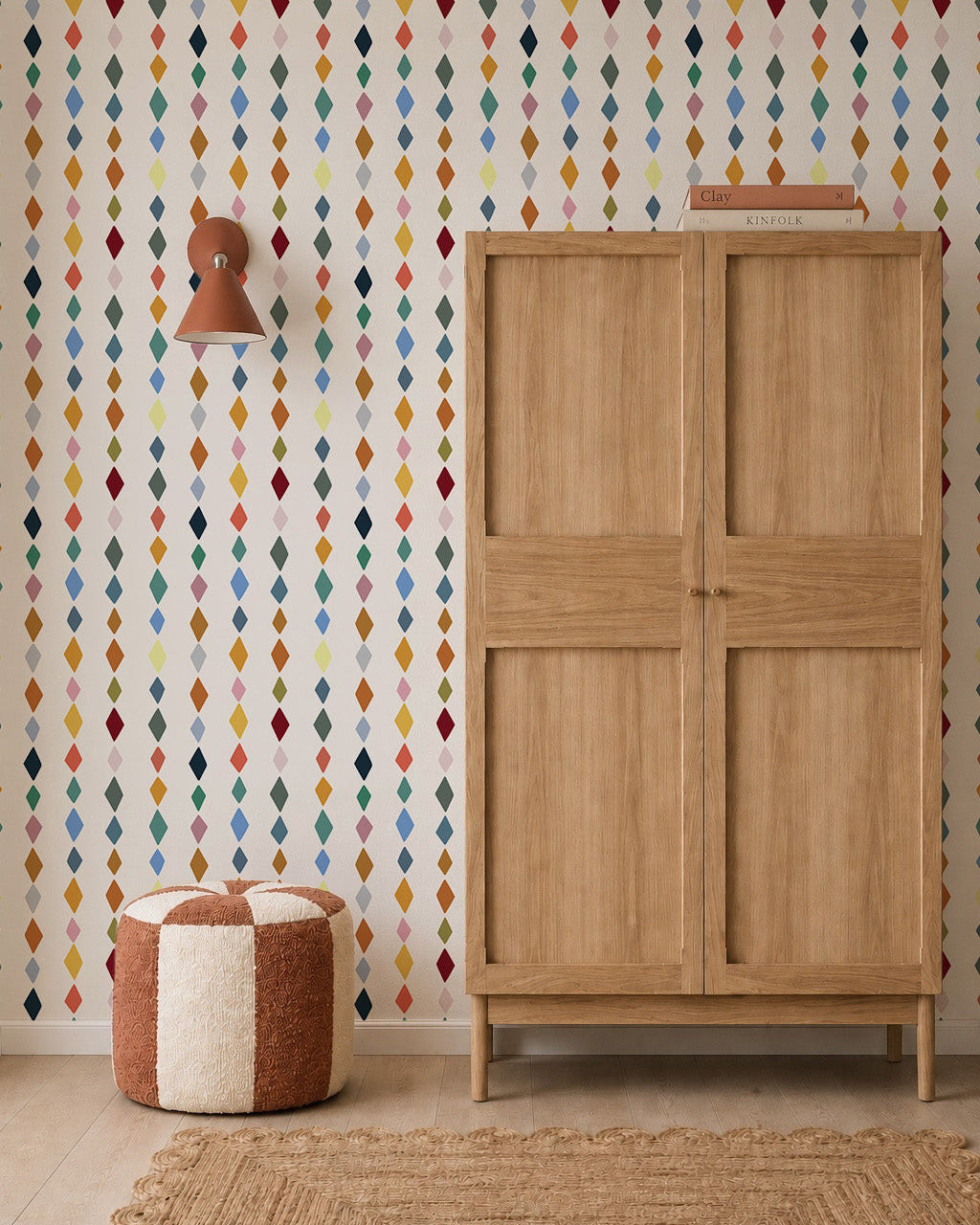 A wooden wardrobe stands next to a brown and white pouf and a wall lamp, against a wall decorated with Jolly Jester Wallpaper, Multi. A woven rug is spread across part of the light wood floor.