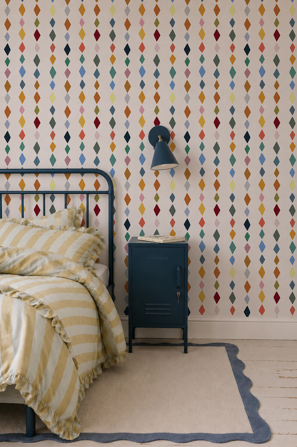 A cozy bedroom features a metal bed frame, yellow and white striped bedding, blue nightstand, wall lamp, painted wooden floorboards with a rug, and walls adorned with Jolly Jester Wallpaper, Multi—colorful diamonds in vertical rows.