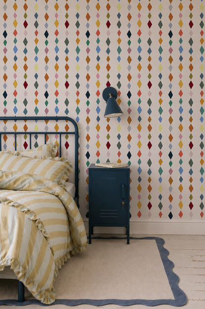 A cozy bedroom features a metal bed frame, yellow and white striped bedding, blue nightstand, wall lamp, painted wooden floorboards with a rug, and walls adorned with Jolly Jester Wallpaper, Multi—colorful diamonds in vertical rows.