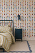 A cozy bedroom features a metal bed frame, yellow and white striped bedding, blue nightstand, wall lamp, painted wooden floorboards with a rug, and walls adorned with Jolly Jester Wallpaper, Multi—colorful diamonds in vertical rows.