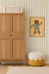 A wooden wardrobe stands beside a round pouf with a white and yellow pattern. Above it hangs a framed picture of a bear in pajamas on a skateboard. The wall features Jolly Jester Wallpaper, Yellow, adding playful charm.