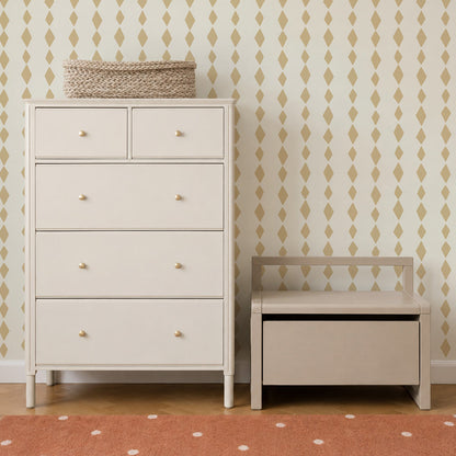 A light beige dresser and a matching storage bench sit against a wall adorned with Jolly Jester Wallpaper in Yellow, featuring a playful gold harlequin pattern. A woven basket rests on top of the dresser, and an orange rug with white dots is partially visible below.