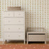 A light beige dresser and a matching storage bench sit against a wall adorned with Jolly Jester Wallpaper in Yellow, featuring a playful gold harlequin pattern. A woven basket rests on top of the dresser, and an orange rug with white dots is partially visible below.