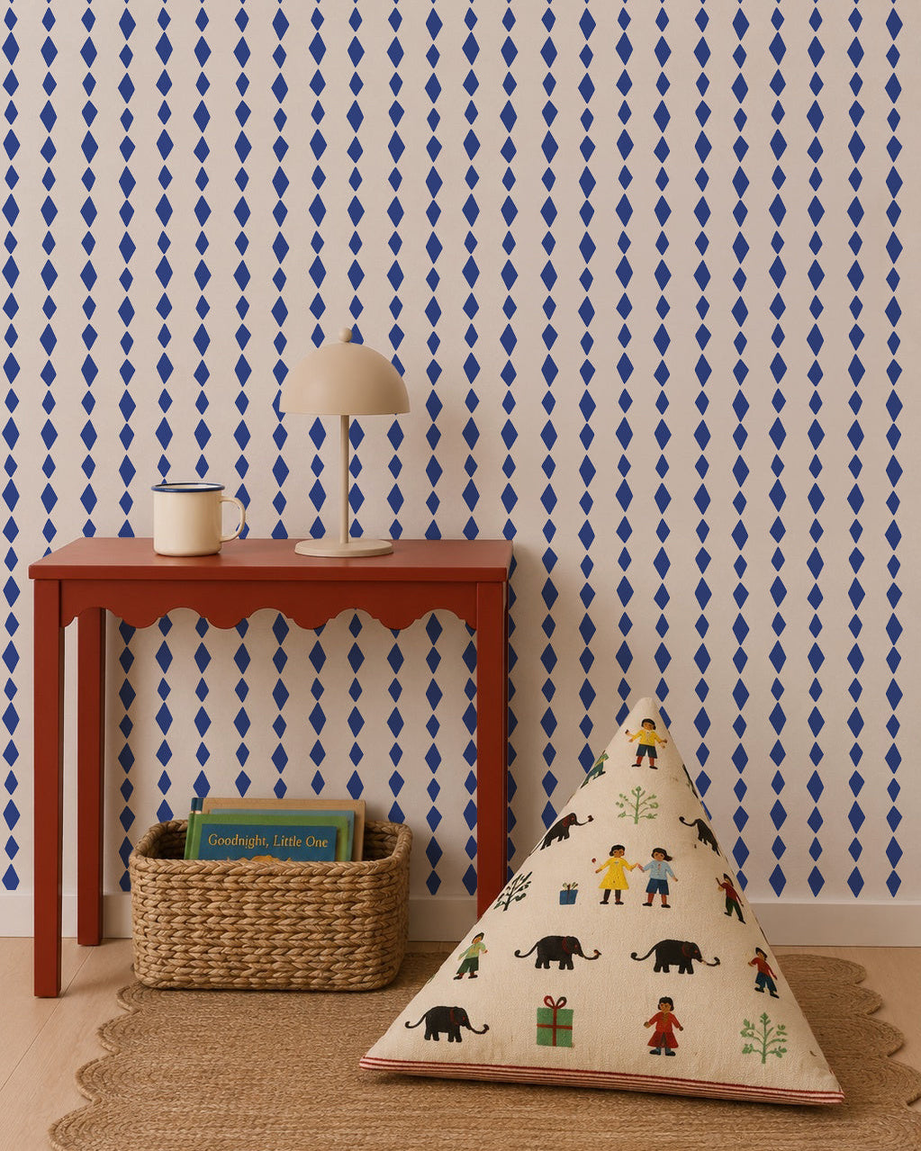 A red table with scalloped edges holds a lamp and mug, with a woven basket of books beneath. A patterned cushion depicting animals and people rests on the floor. The wall showcases Jolly Jester Wallpaper, Royal Blue, in a bold diamond pattern.