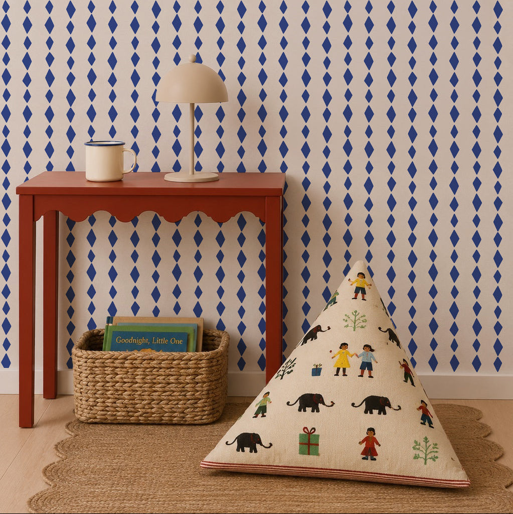 A red table with scalloped edges holds a lamp and mug, with a woven basket of books beneath. A patterned cushion depicting animals and people rests on the floor. The wall showcases Jolly Jester Wallpaper, Royal Blue, in a bold diamond pattern.