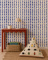 A red table with scalloped edges holds a lamp and mug, with a woven basket of books beneath. A patterned cushion depicting animals and people rests on the floor. The wall showcases Jolly Jester Wallpaper, Royal Blue, in a bold diamond pattern.