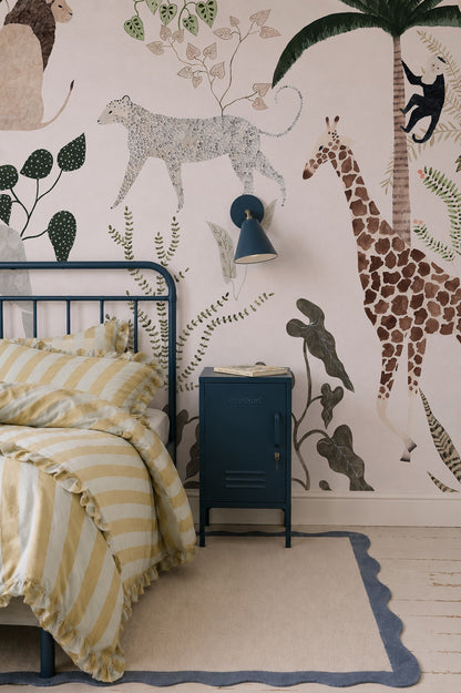 A cozy bedroom featuring the Jungle Dreams Mural with playful giraffes, cheetahs, monkeys, and leafy plants, complemented by a blue bed frame, yellow striped bedding, and a matching blue nightstand.
