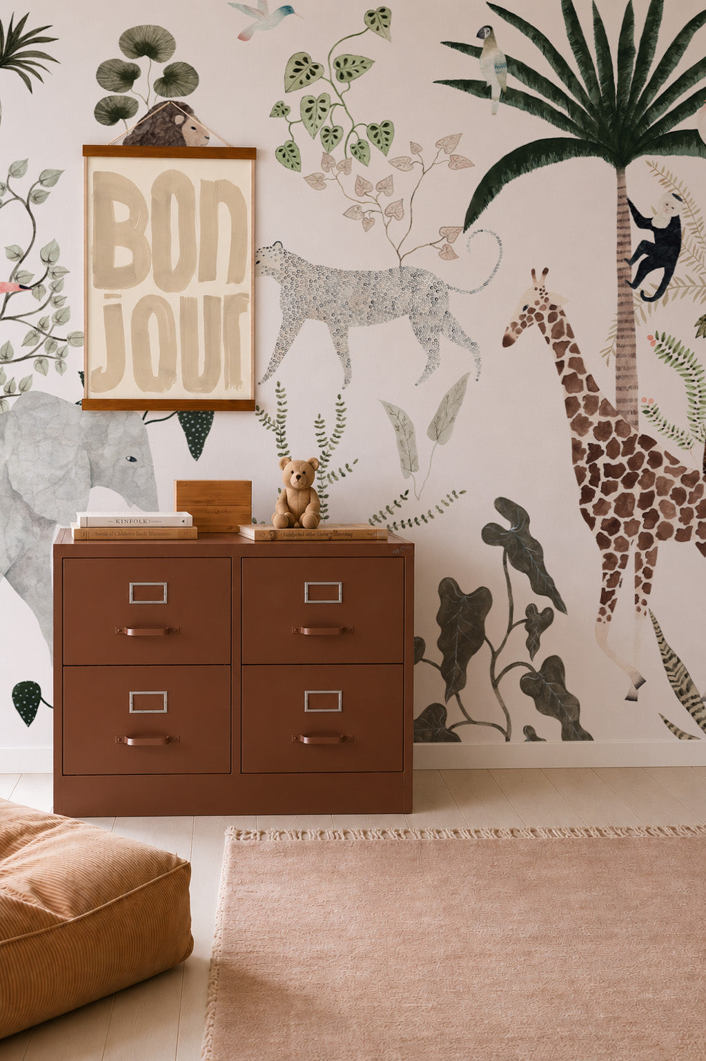 Children’s room with a brown dresser, teddy bear, and the Jungle Dreams Mural showcasing a giraffe, leopard, monkey, and lush plants. A framed Bonjour print hangs above the dresser. Soft tones and playful decor create a cozy atmosphere, further enhanced by the charming Jungle Dreams Mural that brings the nursery to life.