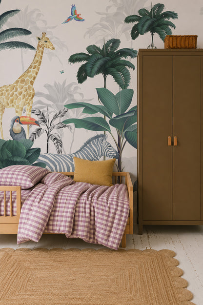 Children’s room with a Jungle Mural featuring safari animals and tropical plants. The bed has a pink gingham duvet, yellow cushion, stands next to a brown wardrobe, and a woven rug is on the floor.