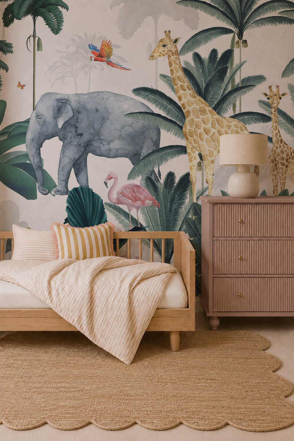 A cozy kids’ room features a wooden bed with striped bedding, a scalloped jute rug, a pink dresser with a lamp, and vibrant Jungle Mural wallpaper with elephants, giraffes, flamingos, and tropical plants.