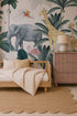 A cozy kids’ room features a wooden bed with striped bedding, a scalloped jute rug, a pink dresser with a lamp, and vibrant Jungle Mural wallpaper with elephants, giraffes, flamingos, and tropical plants.