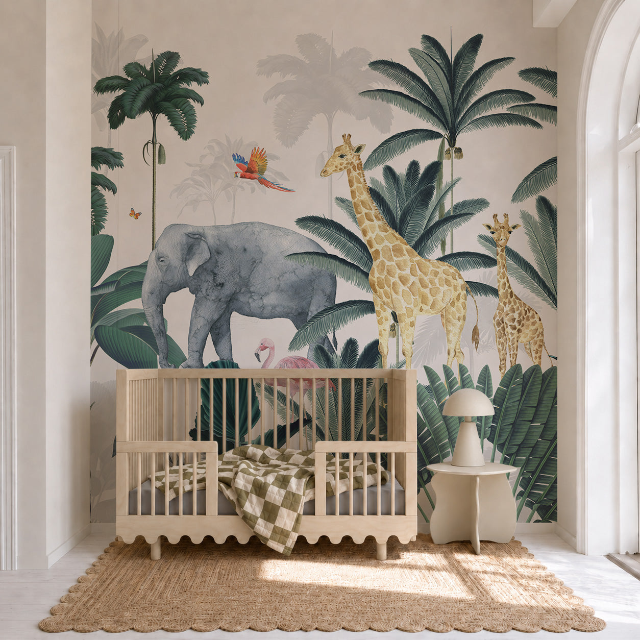 A nursery featuring a wooden crib, checkered blanket, small table lamp, and a vibrant Jungle Mural with safari animals like an elephant, giraffes, a flamingo, palm trees, green leaves, and a parrot. Natural light floods the charming kids’ room through a large arched window.