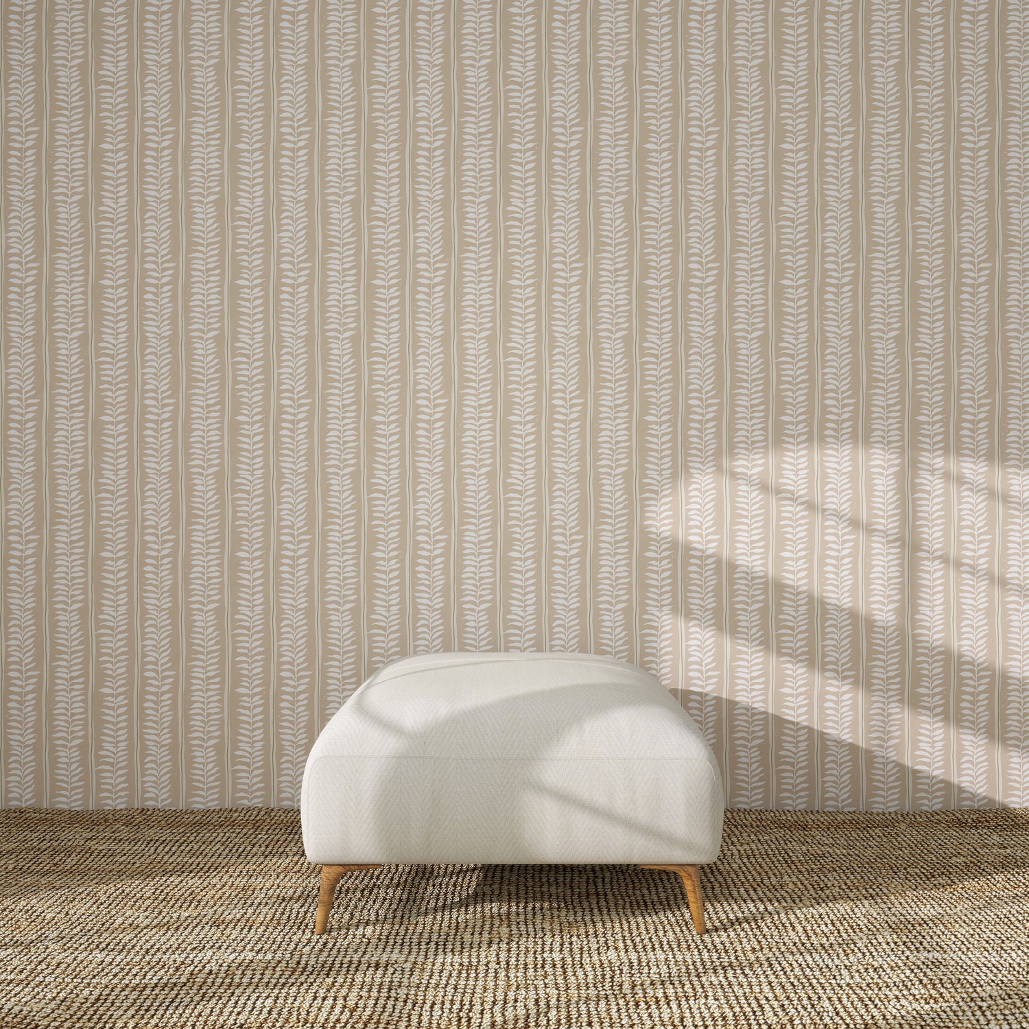A white upholstered ottoman with wooden legs rests on a textured brown rug before Meadow Leaf Stripe Wallpaper in Clay, as sunlight highlights the botanical charm of the leaf-patterned backdrop.