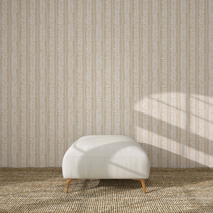 A white upholstered ottoman with wooden legs rests on a textured brown rug before Meadow Leaf Stripe Wallpaper in Clay, as sunlight highlights the botanical charm of the leaf-patterned backdrop.