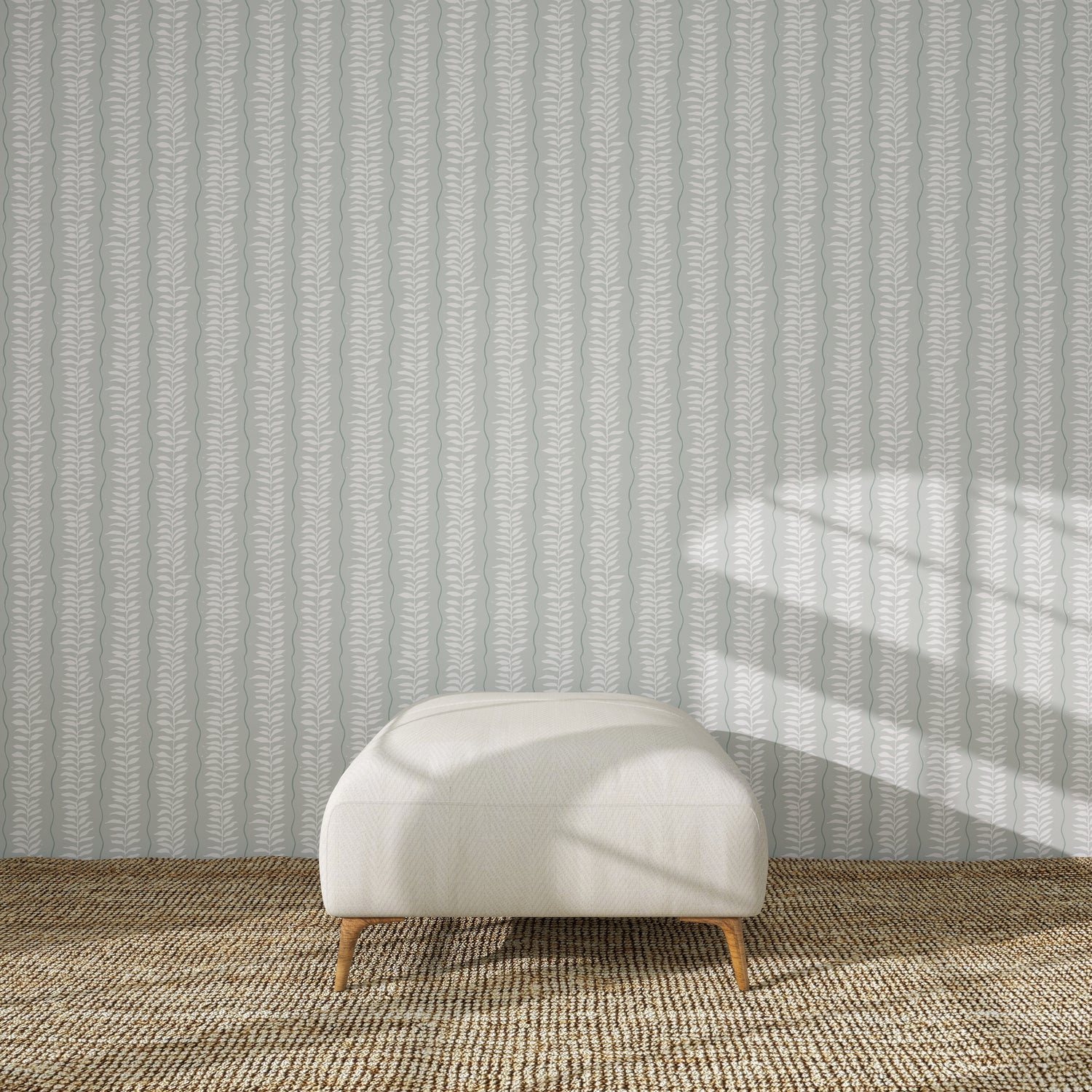 A white upholstered ottoman with wooden legs sits on a woven beige rug in front of the Meadow Leaf Stripe Wallpaper Sample in Sea Foam. Sunlight casts window-shaped shadows, highlighting the ottoman’s color and quality.