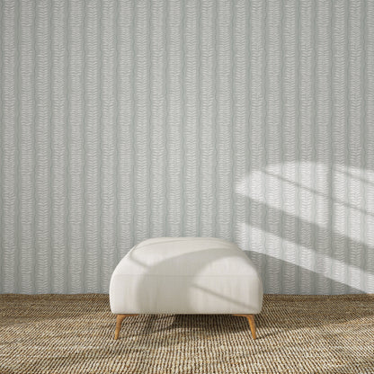 A white upholstered ottoman with wooden legs sits on a woven beige rug in front of the Meadow Leaf Stripe Wallpaper Sample in Sea Foam. Sunlight casts window-shaped shadows, highlighting the ottoman’s color and quality.